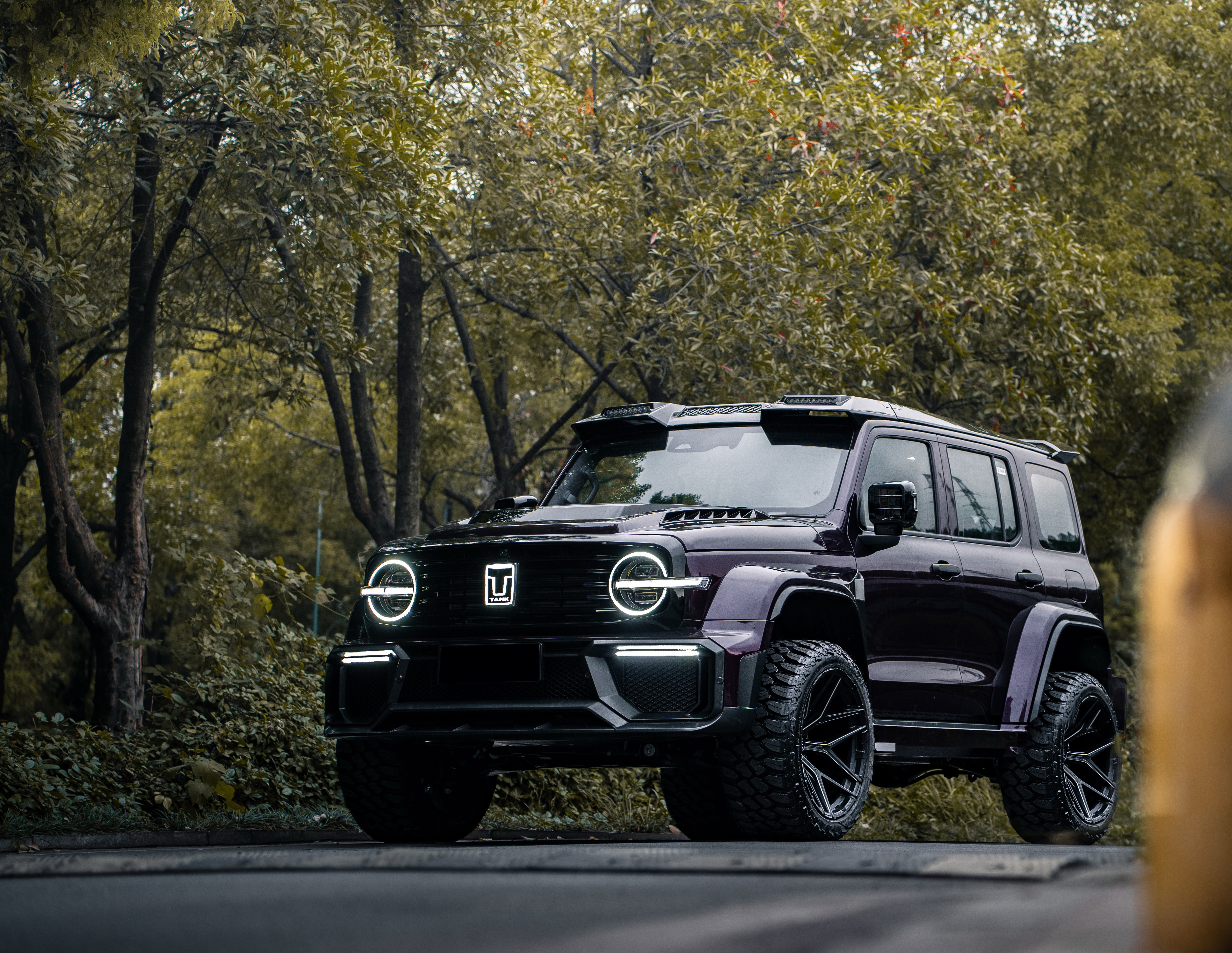 Black off-road SUV with illuminated round headlights parked on a forest road surrounded by green trees.